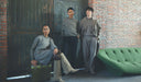 Three people in matching gray outfits standing against a brick wall with a green sofa.