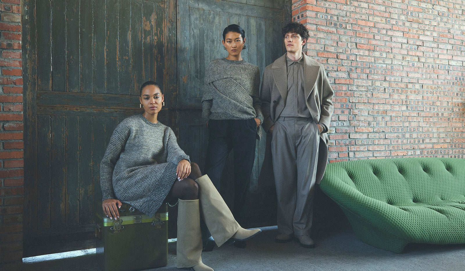 Three people in matching gray outfits standing against a brick wall with a green sofa.