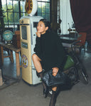 Woman in a black outfit sitting on a motorcycle next to an old-fashioned phone booth.