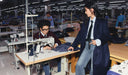Person using a sewing machine in a factory setting with another person observing.