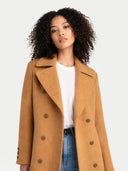 Woman wearing a tan double breasted coat over a white shirt and blue jeans on a light gray background  #color_almond