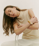 Woman wearing a beige sleeveless top and white pants against a plain background