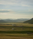 Scenic view of a vast grassland with hills and a few structures under a cloudy sky.