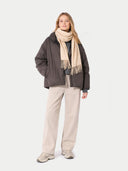 Woman wearing a dark jacket, beige scarf, and light pants on a white background