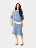 Woman wearing a blue sweater and skirt with a white shirt underneath, standing against a light gray background. #color_dusty-blue