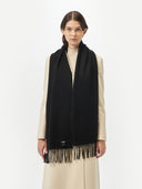 front view of a woman wearing a black oversized woven scarf with fringe over a beige long coat in a light gray background.