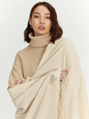 Woman wearing a beige turtleneck sweater and holding a cream-colored blanket with fringe on a white background