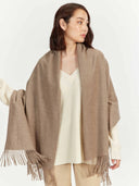Woman wearing a beige scarf with fringe over a white sweater and beige pants on a white background