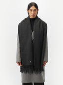 front view of a man wearing an oversized cashmere woven scarf in dark gray color over a gray coat in a light gray background.