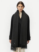front view of a woman wearing an oversized cashmere woven scarf in dark gray color over a black coat in a light gray background.