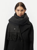 close up front view of a woman wearing an oversized cashmere woven scarf in dark gray color over a black coat in a light gray background.
