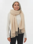 front view of a woman wearing a white sweater with wrapped oversized beige scarf with fringe and black pants.