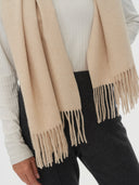 close up of a woman standing with a white long sleeve top, beige scarf with fringe and black pants.