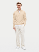 A man modeling a light beige quarter-zip knit sweater over a white t-shirt, paired with off-white trousers and white sneakers, against a solid light gray background.