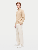 A man modeling a light beige quarter-zip knit sweater, paired with off-white trousers and white sneakers, against a solid light gray studio background.