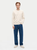 A man wearing a cream crewneck sweater, blue jeans, and off-white sneakers, standing against a light gray studio background.