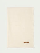 A cream-colored woven scarf with a small brown leather brand patch on the bottom right corner, displayed flat against a light off-white background.