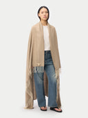 A model stands against a light gray studio background wearing a long beige fringed wrap or shawl over a white top and blue wide-leg jeans.