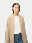 A woman modeling a beige textured knit wrap or shawl over a white t-shirt and blue jeans, shot against a solid light gray background.