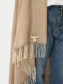 A close-up of a beige and cream herringbone patterned scarf with fringe, draped over blue jeans against a light gray background.