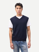 Man wearing a navy sleeveless sweater over a white shirt with blue jeans on a light gray background. #color_navy-blazer