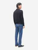 A man shown from the back wearing a black mock-neck knit sweater and blue jeans, standing against a plain light gray studio background.