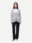 A woman modeling a light gray knit cardigan with dark buttons over a white top and black wide-leg pants, standing against a plain light gray background.