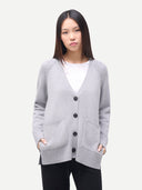 A woman modeling a light gray knit cardigan with black buttons and front pockets, worn over a white top and black pants, against a plain light gray background.