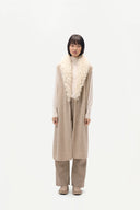 A model stands against a plain white studio background wearing a long beige knit vest with a thick cream boucle collar, a cream turtleneck, beige wide-leg trousers, and beige shoes.