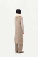 A model viewed from the back wearing a long beige textured knit vest with a fluffy cream collar, layered over a light beige long-sleeve top and wide-leg beige trousers, against a solid white background.