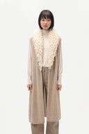A model stands against a plain white studio background wearing a long beige knit vest with a large cream bouclé collar over a cream turtleneck and beige trousers.