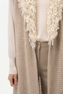 A close-up of a person wearing a long cream knit cardigan with a chunky shaggy fringe trim over a cream top and taupe pants.