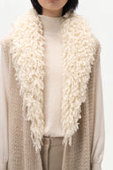 A close-up of a person wearing a chunky cream shaggy knit scarf layered over a beige ribbed knit vest, cream turtleneck, and beige trousers against a plain white background.