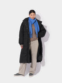 front view of a woman wearing a black long puffer jacket, blue fringed scarf, brown sweater, gray pants on a light gray background #color_blue