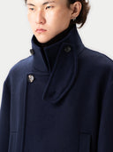 Close-up of a person wearing a navy blue coat with a high funnel neck and a buttoned throat latch overlay. The coat features large dark buttons and is layered over a dark turtleneck #color_navy-blazer