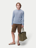 Man wearing a light blue sweater and brown shorts, holding a green bag on a white background. #color_celestial-blue