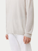 A close-up shot of a person wearing a light oatmeal-colored, fine-knit sweater with ribbed cuffs and a rolled hem, paired with white trousers.  #color_silver-birch