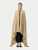 front view of a person wrapped in a beige cable knit blanket with a subtle pattern and black skirt on a light gray background #color_beige