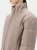 A close-up side view of a person wearing a taupe, quilted puffer jacket with a high collar and a front zipper, set against a plain light gray background. #color_taupe