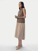 A model is shown from the side wearing a sleeveless taupe-brown ribbed knit mock-neck sweater vest with matching ribbed knit arm warmers, paired with a pleated beige midi skirt and beige pointed-toe flats, against a light gray studio background.