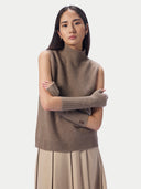 A model wearing a taupe brown sleeveless mock-neck knit sweater vest with matching ribbed knit arm warmers, paired with a pleated beige skirt, against a light gray studio background.