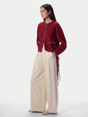 A model is shown from the side wearing a deep red cropped cardigan with decorative chain-like trim and fringe details, paired with wide-leg cream trousers, against a plain light gray studio background.