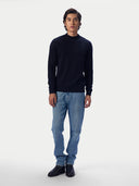 A man modeling a dark navy blue mock neck knit sweater, paired with light blue jeans and black boots, standing against a plain light gray studio background.