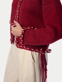 A close-up of a person wearing a deep red cropped knit cardigan with braided trim and tassel ties, paired with light beige trousers, against a plain light background.
