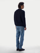A man wearing a dark navy blue knit sweater and light blue jeans, shown from a rear three-quarter view against a plain light gray studio background.