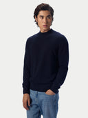 A man wearing a dark navy blue mock neck knit sweater and light blue jeans, standing against a light gray studio background.