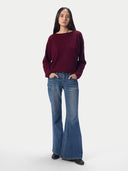 A woman modeling a long-sleeve boat-neck knit sweater in deep burgundy, paired with blue flare jeans and black shoes, against a plain light gray studio background.