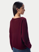 A woman shown from the back wearing a loose-fitting, long-sleeve knit sweater in deep burgundy, paired with blue jeans, against a plain light gray studio background.