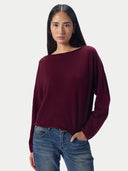 A woman modeling a loose, boat-neck knit sweater in deep burgundy with blue jeans, set against a plain light gray studio background.