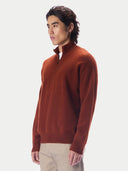 A man wearing a rust-brown quarter-zip knit sweater and beige trousers, photographed against a clean light gray studio background.